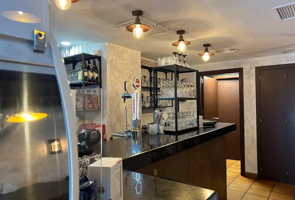 Bar counter with a black top and a beer tap at Bar El Pozo on Plaza de la Corredera in Córdoba, shelves of glasses and bottles under copper pendant lights