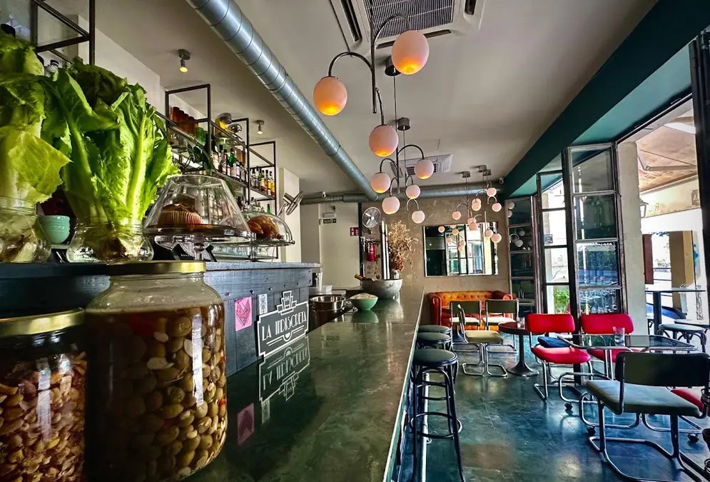 Interior of La Indiscreta bar in Córdoba's Ribera with a long green marble counter, glass cake domes, jars of olives, pendant globe lights, red chrome chairs and open glass doors to the street